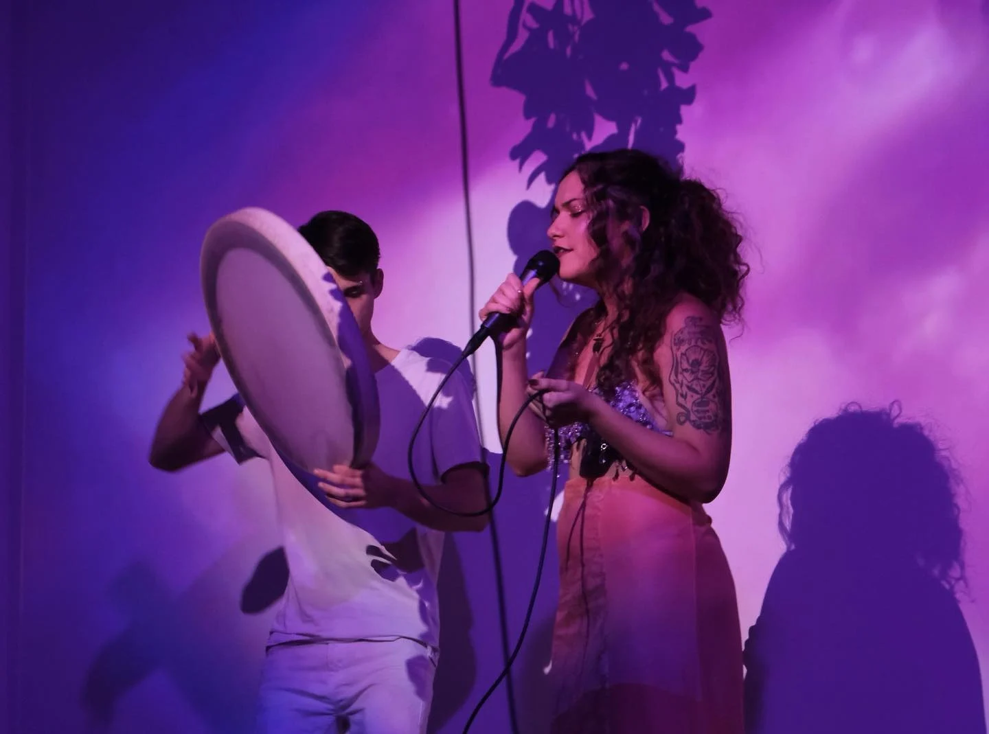 Vocalist singing into a microphone beside a frame-drum player under stage light