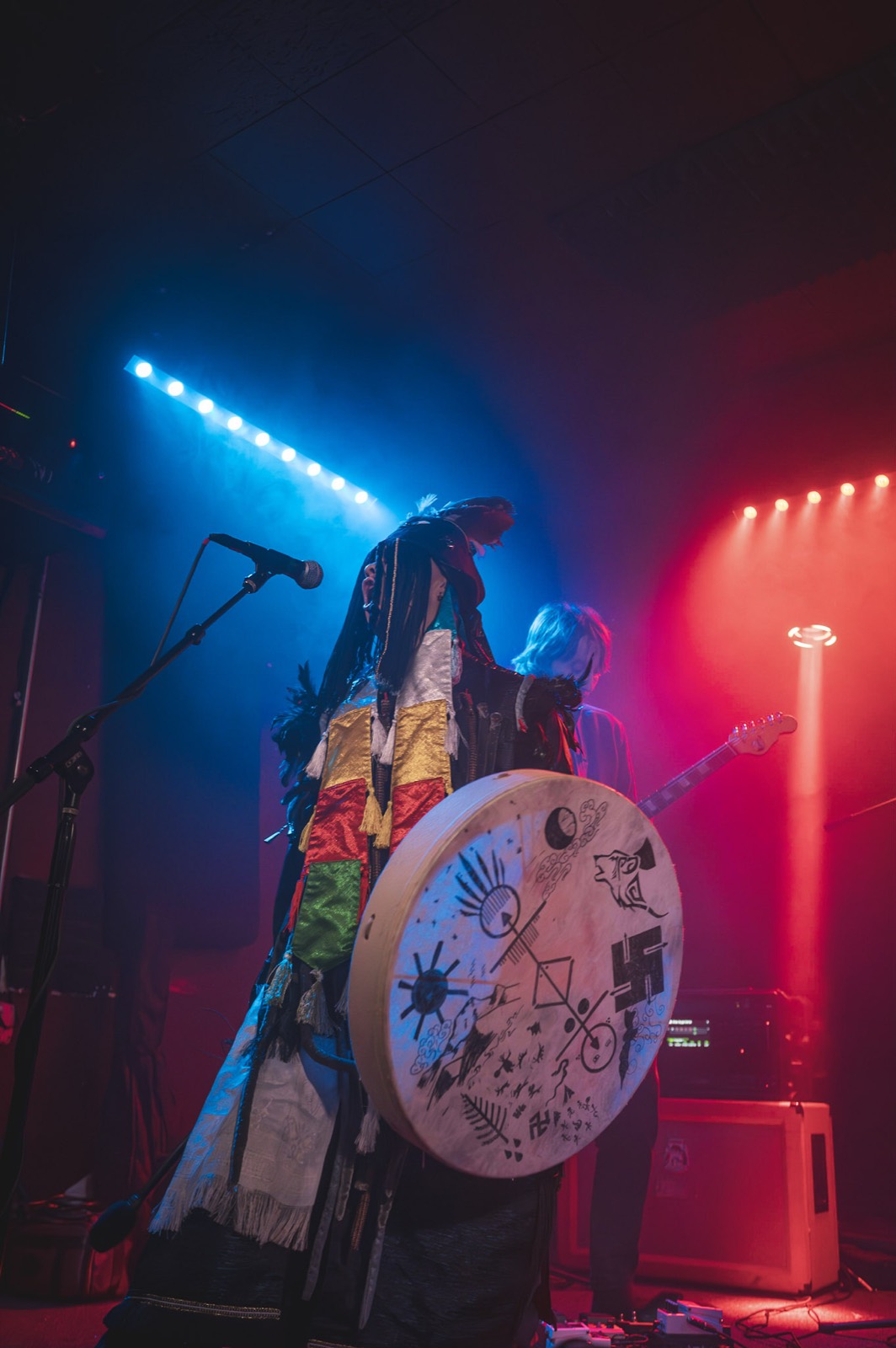 Performer in shamanic dress at a microphone holding a painted frame drum